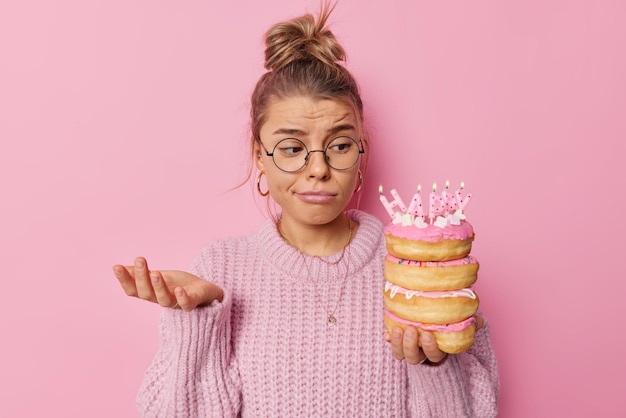 Mulher bonita e triste questionada levanta a palma da mão parece descontente com deliciosos donuts com velas acesas se sente infeliz quando comemora o aniversário sozinho usa óculos redondos e suéter de malha