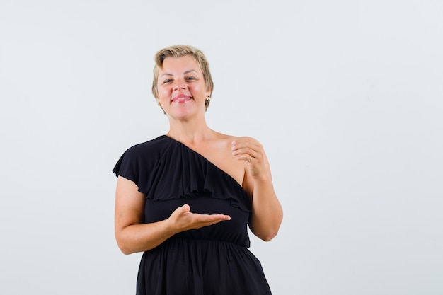 Foto grátis mulher bonita em blusa preta espalhando a palma da mão aberta, mostrando algo e parecendo satisfeita