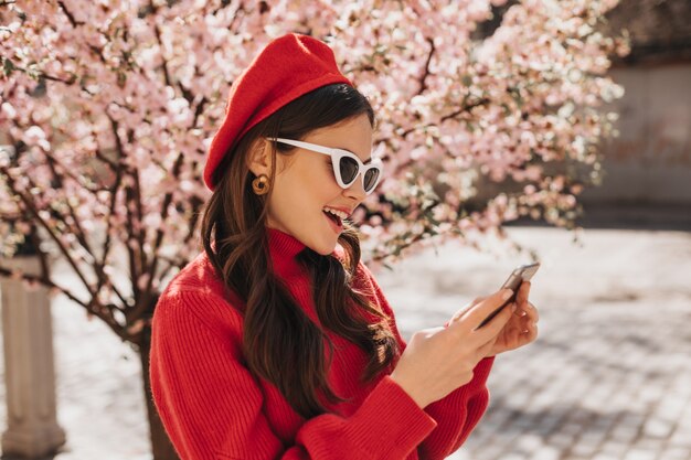 Mulher bonita em boina e óculos de sol está conversando no telefone perto de Sakura. Retrato externo de senhora com suéter vermelho cashemere segurando um celular