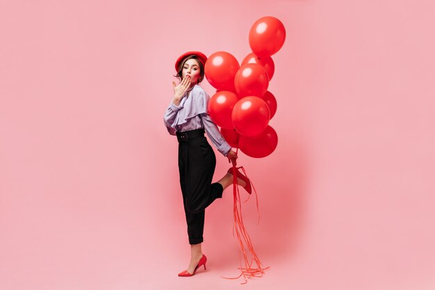 Mulher bonita em calças pretas elegantes, blusa e boina brilhante sopra beijo, provocantemente levanta a perna e segura balões no fundo rosa.