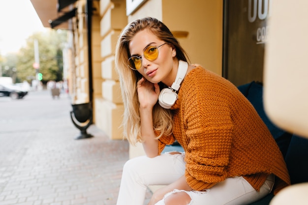 Mulher bonita em jeans branco e casaco de lã marrom olhando com um sorriso interessado enquanto descansa no banco