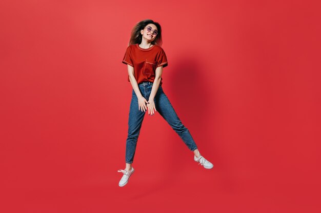 Mulher bonita em jeans e camiseta brilhante pulando na parede vermelha
