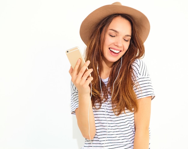 Mulher bonita em roupas de verão casual hipster sem maquiagem no chapéu marrom, ouvindo música no telefone com fones de ouvido isolados no branco