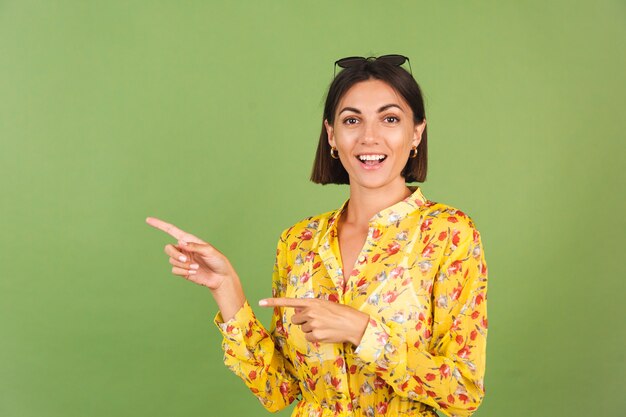 Mulher bonita em um vestido amarelo de verão e óculos escuros, estúdio verde, animada alegre alegre apontar o dedo esquerdo