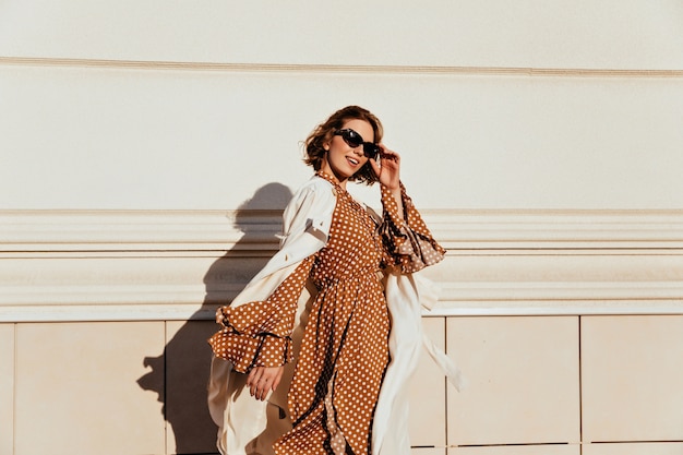 Mulher bonita em um vestido marrom longo, aproveitando o dia de sol. adorável garota branca em traje retrô, andando pela rua.