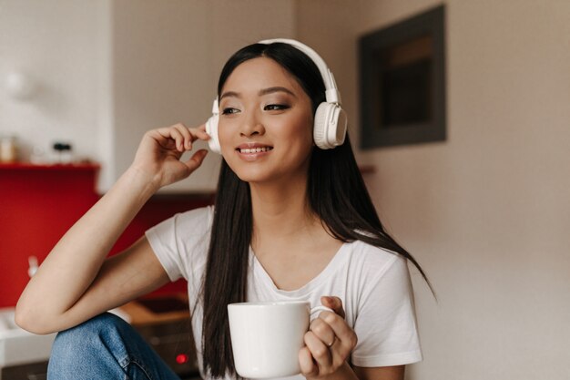 Mulher bonita em uma camiseta branca e fones de ouvido está ouvindo música enquanto toma uma xícara de chá