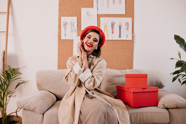Mulher bonita em uma trincheira na moda e poses de boina de estilo francês no apartamento. linda garota com boina vermelha e casaco bege outono está sorrindo.