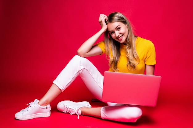 Mulher bonita encaracolada jovem feliz sentada no chão com as pernas cruzadas e usando o laptop na parede vermelha.