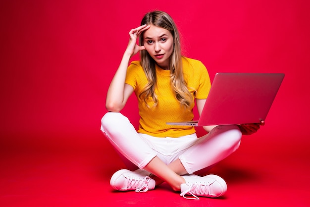 Mulher bonita encaracolada jovem feliz sentada no chão com as pernas cruzadas e usando o laptop na parede vermelha.