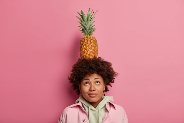 Mulher bonita encaracolada olha para cima, mantém o equilíbrio com o abacaxi na cabeça, concentrada acima, tem pele morena e cabelos encaracolados, vestida casualmente, isolada na parede rosa. linda fêmea trazendo frutas tropicais