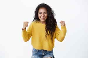 Foto grátis mulher bonita encorajada e motivada em um suéter amarelo levantando as mãos, soco de felicidade, sorrindo, ouvir boas notícias, comemorando a vitória, ganhando uma grande aposta, parede branca