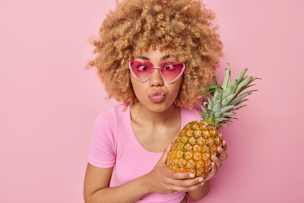 Mulher bonita engraçada usa óculos de sol em forma de coração faz careta na câmera mantém os lábios cruzados olhos cruzados segura abacaxi maduro fresco isolado sobre fundo rosa Frutas e vitaminas saudáveis