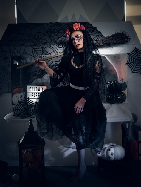 Mulher bonita está comemorando o Halloween de vestido preto e maquiagem incomum enquanto segura a vassoura.