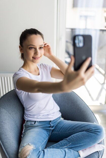 Mulher bonita fazendo selfie com seu telefone inteligente e sorrindo enquanto está sentada em uma poltrona em casa
