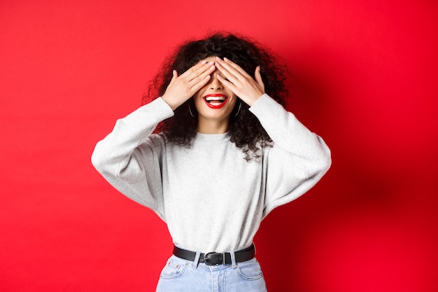 Mulher bonita feliz com cabelos cacheados e lábios vermelhos, cobrindo os olhos com as mãos e esperando surpresa, sorrindo animado, em pé contra um fundo vermelho.