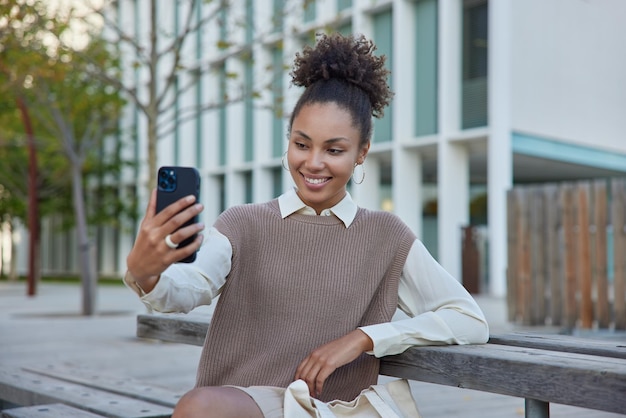 Mulher bonita feliz com cabelos cacheados reunidos em coque grava vídeo de selfie ou poses para foto usa smartphone vestido com roupas elegantes posa ao ar livre em ambiente urbano Conceito de estilo de vida moderno