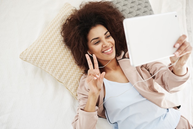 Mulher bonita feliz em fones de ouvido sorrindo mostrando paz fazendo selfie tablet deitado na cama. de cima.