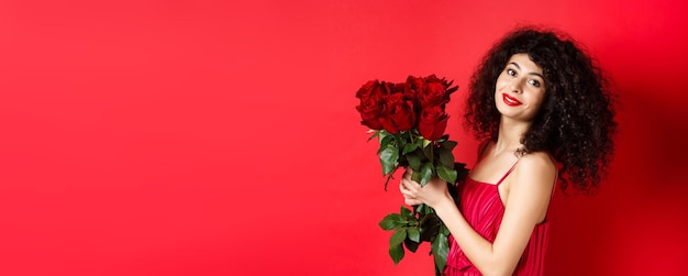 Mulher bonita feliz vestido segurando flores e sorrindo romântico em pé contra o fundo vermelho