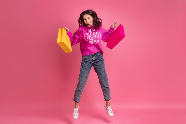 Mulher bonita hispânica com capuz rosa e jeans sorrindo pulando no rosa isolada segurando sacolas coloridas com desconto na venda