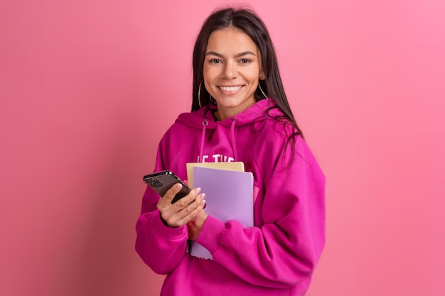 Mulher bonita hispânica com capuz rosa sorrindo segurando notebooks e usando smartphone posando em fundo rosa isolado