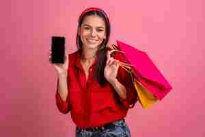 Foto grátis mulher bonita hispânica de camisa vermelha sorrindo segurando sacolas de compras e smartphone em fundo rosa estúdio isolado venda de desconto sexta-feira negra