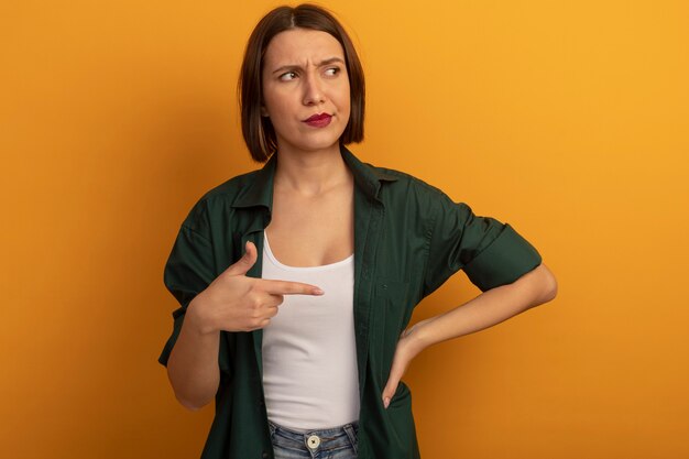 Mulher bonita irritada olha e aponta para o lado isolado na parede laranja