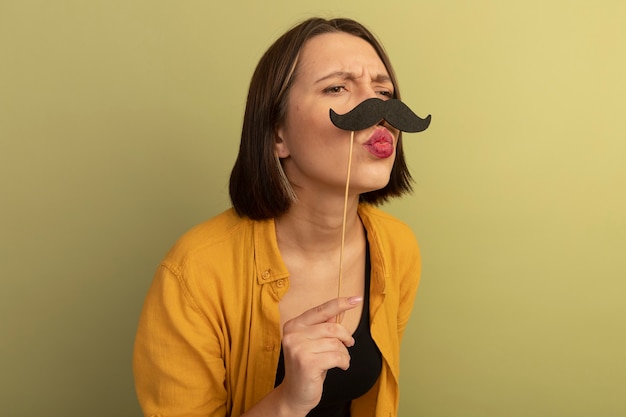 Foto grátis mulher bonita irritada segurando bigode falso em uma vara olhando para o lado isolado na parede verde oliva