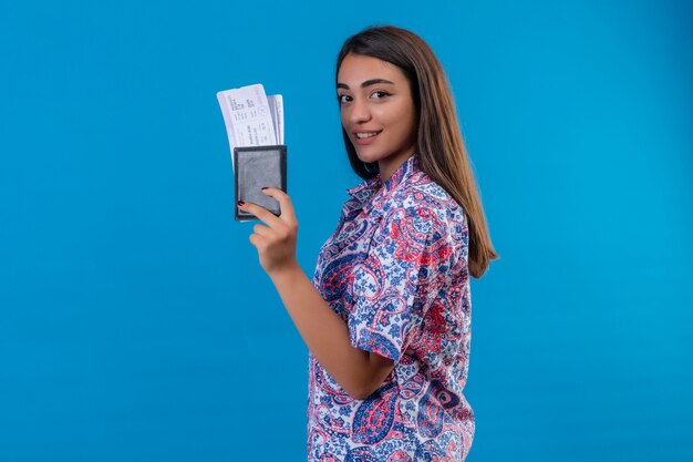 Mulher bonita jovem satisfeita com bilhetes e passaporte, olhando confiante sorrindo em pé sobre a parede azul