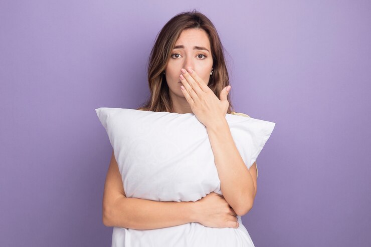 Foto grátis mulher bonita jovem triste em uma camiseta bege segurando um travesseiro branco, olhando para a câmera preocupada, cobrindo a boca com a mão em pé sobre um fundo roxo