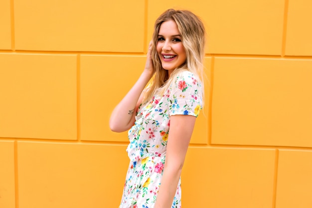 Mulher bonita loira com vestido floral, posando perto de uma parede amarela