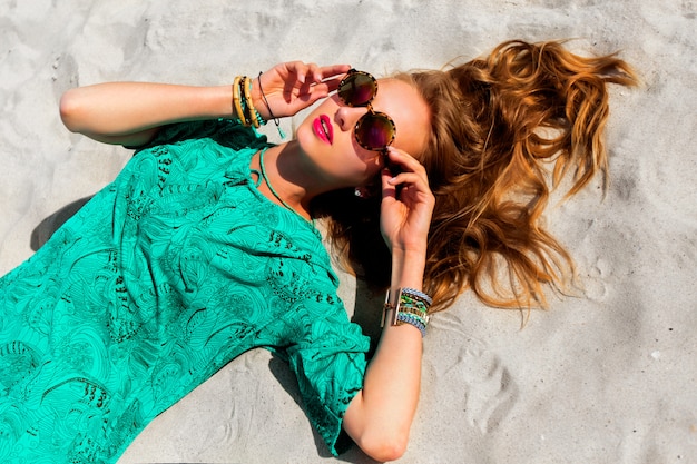 Mulher bonita loira deitada em uma praia tropical ensolarada, usando óculos escuros elegantes e legais, túnica boho colorida e acessórios brilhantes da moda