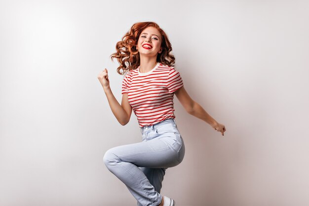 Mulher bonita magro em jeans dançando. Menina sorridente afável pulando na parede branca.