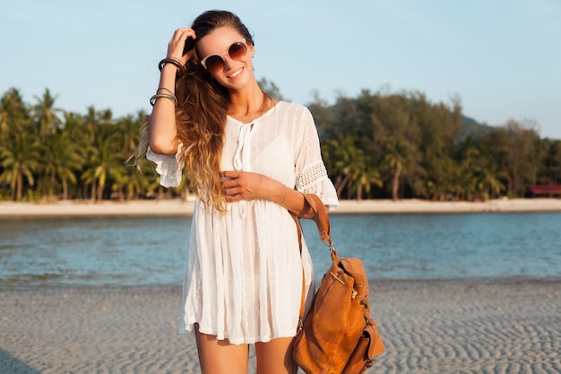 Mulher bonita magro em um vestido branco na praia tropical no pôr do sol segurando a mochila de couro.