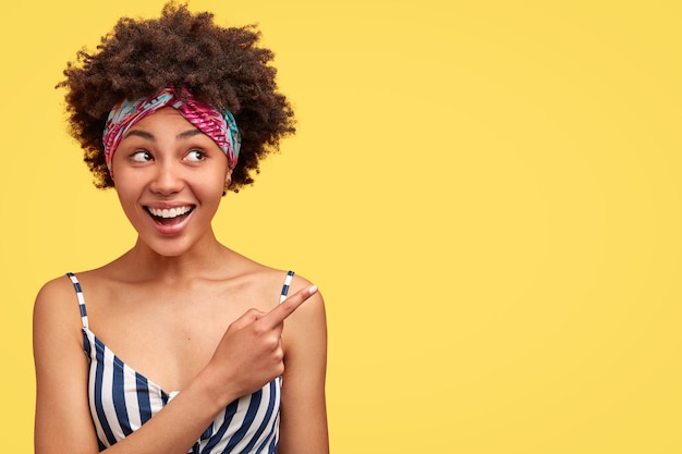 Mulher bonita mestiça com cabelo crespo, sorriso gentil, mostra algo agradável, indica com o dedo indicador na parede amarela em branco. Mulher afro-americana encantadora posando em ambientes internos