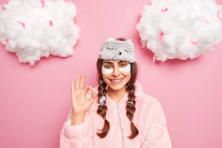 Foto grátis mulher bonita morena caucasiana sorri gentilmente mostra gesto de ok gosta de algo aplica manchas de colágeno para reduzir o inchaço sob os olhos, vestida com pijamas macios isolados sobre a parede rosa