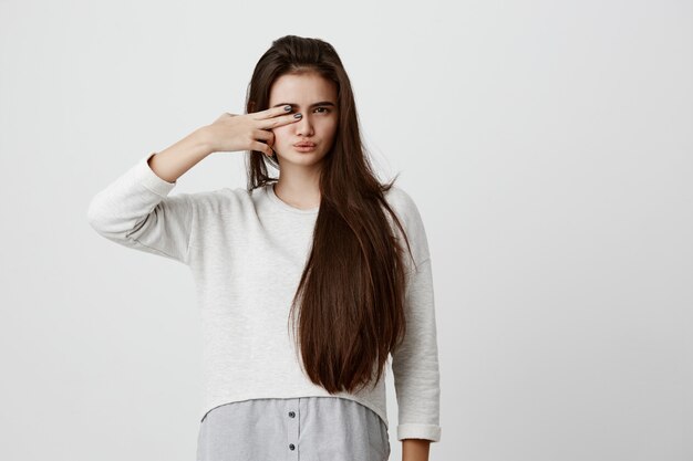 Mulher bonita morena com cabelos longos, fazendo caretas, fazendo beicinho os lábios, fechando um dos olhos com sinal de v, tendo expressão agradada. Linda fêmea jovem vestida com roupas casuais, posando dentro de casa