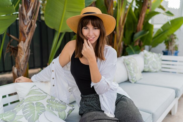Mulher bonita morena com chapéu de palha sentada em um aconchegante terraço tropical