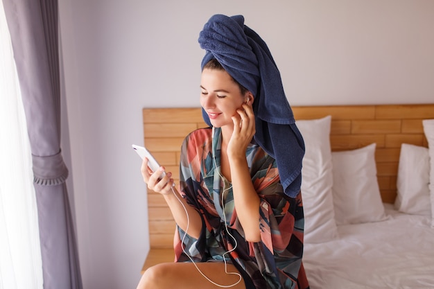 Mulher bonita morena, envolvida em uma toalha de banho usando telefone celular enquanto está sentado na cama, ouvindo música por fones de ouvido