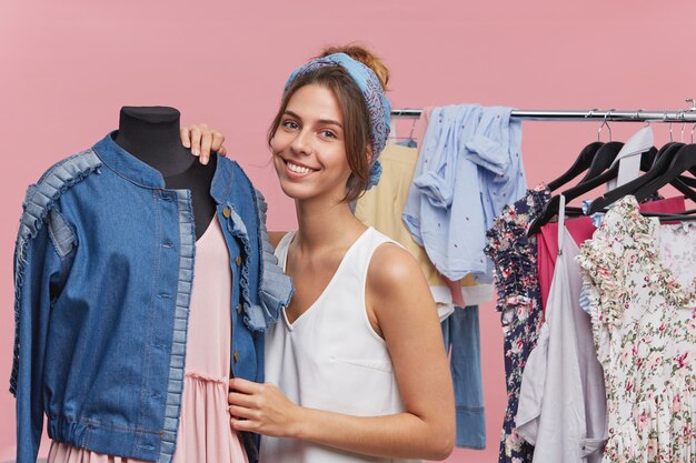 Mulher bonita morena vestindo camiseta branca e lenço na cabeça, em pé perto de manequim com jaqueta jeans e vestido rosa, em pé no provador, de bom humor. Selecionando roupas para festa