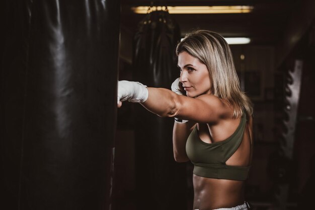 Mulher bonita musculosa está fazendo seus exercícios de kickboxing com saco de pancadas.