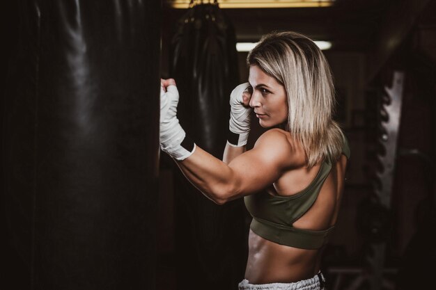 Mulher bonita musculosa está fazendo seus exercícios de kickboxing com saco de pancadas.