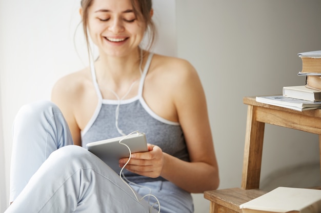 Mulher bonita nova nos fones de ouvido que sorri olhando a tabuleta que escuta fluindo a música que senta-se no assoalho sobre a parede branca.