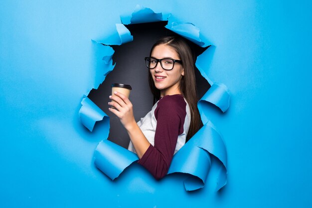 Mulher bonita nova que guarda o café ao olhar através do furo azul na parede de papel.