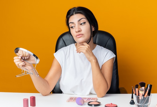 Mulher bonita pensando se senta à mesa com ferramentas de maquiagem segurando e olhando no espelho colocando a mão no queixo