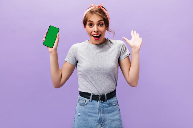 Mulher bonita posa alegremente com smartphone. Jovem alegre em t-shirt cinza elegante e saia jeans posando em fundo isolado.