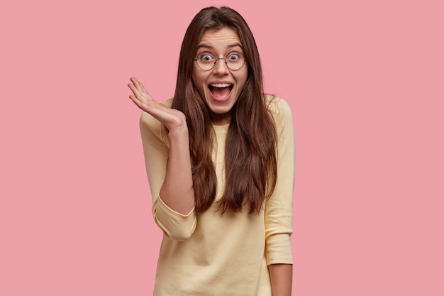 Mulher bonita radiante mantém a mão levantada, exclama de felicidade, tem cabelo comprido, vestida com roupa casual, isolada sobre o espaço rosa