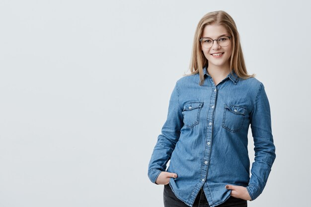 Mulher bonita satisfeita com cabelo loiro liso, olhos escuros, óculos elegantes e pele saudável, vestida com camisa jeans, de mãos dadas nos bolsos sorrindo. Pessoas e estilo de vida