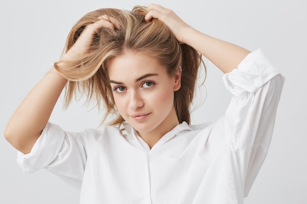 Mulher bonita satisfeita com cabelos loiros, olhos escuros e pele saudável, vestida com camisa branca, segurando as mãos nos cabelos enquanto posava contra a parede de concreto cinza. Conceito de pessoas e beleza.