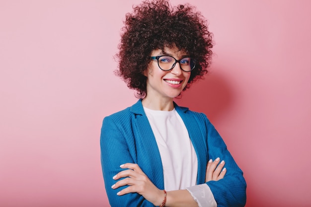 Foto grátis mulher bonita sorridente com cabelo curto e encaracolado vestida de jaqueta e óculos posa rosa com as mãos entrelaçadas e um sorriso maravilhoso