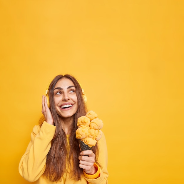 Foto grátis mulher bonita sorridente com uma expressão alegre focada acima segurando um saboroso sorvete de casquinha e pensando em algo agradável enquanto ouve música através de fones de ouvido poses contra a parede amarela